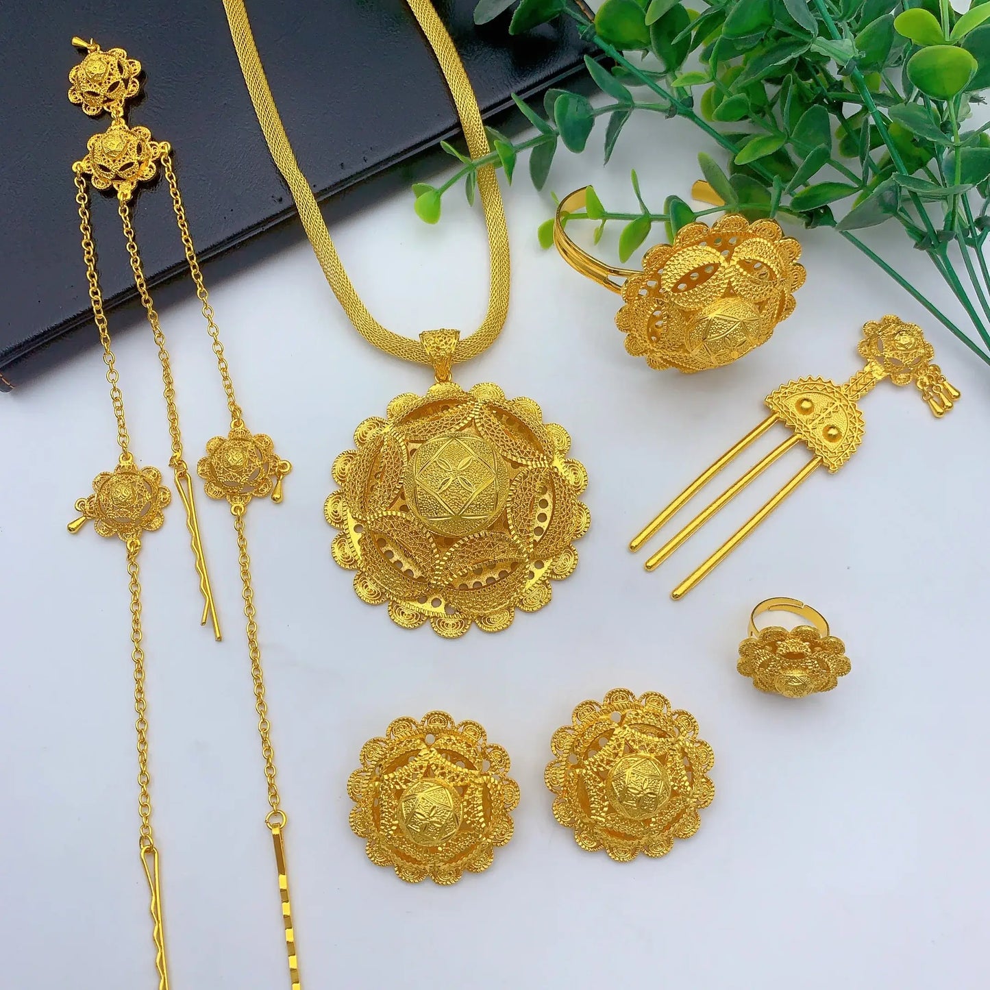 Gold jewelry set including necklace, ring, earrings, and hairpin on a white surface with green leaves.